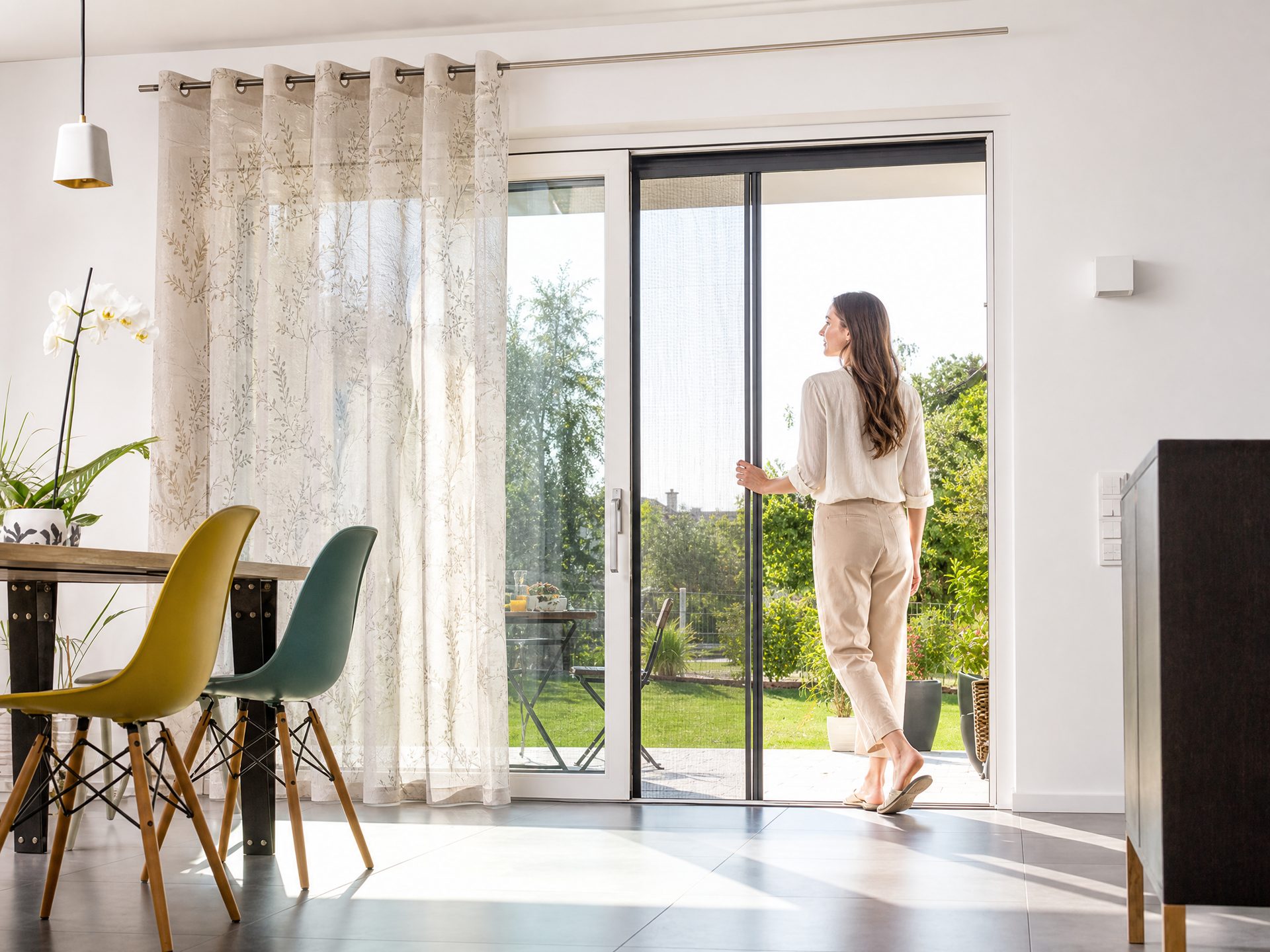 Frau &ouml;ffnet eine Terrassent&uuml;r mit Insektenschutz, dahinter Gardinen im Wohnzimmer